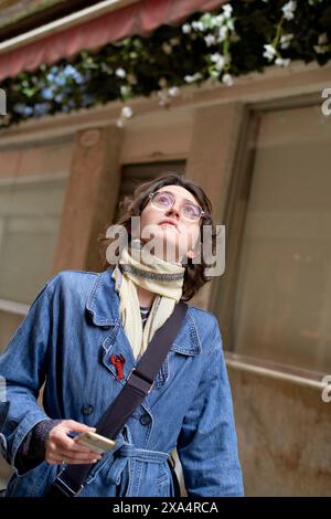 Curious woman with glasses looking upwards holding a smartphone on a city street. Stock Photo