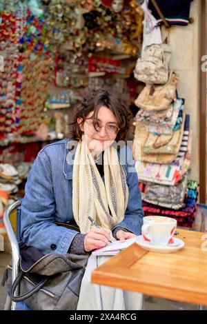 Smiling woman writing in a notebook at an outdoor cafe with colorful accessories in the background. Stock Photo