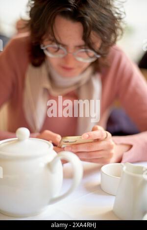Young woman intently checking her smartphone at a cozy cafe table with a teapot in the foreground. Stock Photo