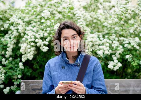 Woman with curly hair using smartphone in a garden with white blossoms. Stock Photo