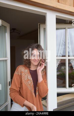 Smiling woman with glasses standing by an open door with curtains in the background. Stock Photo