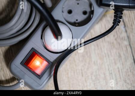 Close-up of a power strip with a red illuminated switch and multiple plugs connected. The power strip is placed on a wooden surface. Stock Photo