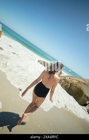 A woman in a black swimsuit walks towards the sea on a sunny beach with waves crashing on rocks. Stock Photo