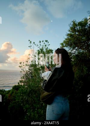A woman takes a photo with her smartphone at sunset by the sea surrounded by greenery. Stock Photo