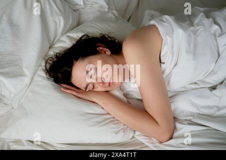 A woman sleeping peacefully in a white bed with her head resting on a pillow, her dark hair spread out, and a serene expression on her face. Stock Photo