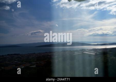 Sunlight streaming through clouds over a coastal town with a clear view of a calm sea and distant hills. Stock Photo