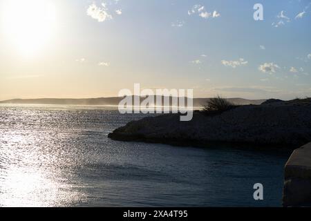 Sunset casting a warm glow over a tranquil sea with distant mountains and a rocky shoreline. Stock Photo