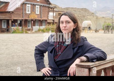 Confident woman leaning on a wooden fence in a western-style town setting with a thoughtful expression. Stock Photo