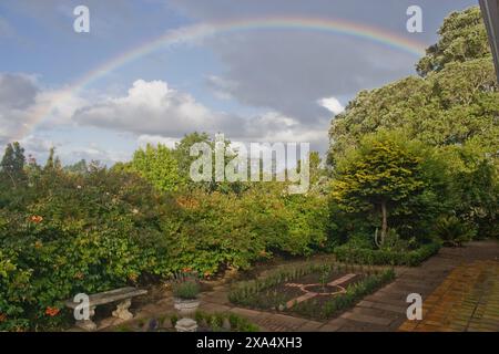 Vibrant rainbow arching over a serene garden with lush greenery and a clear blue sky after rainfall. Stock Photo