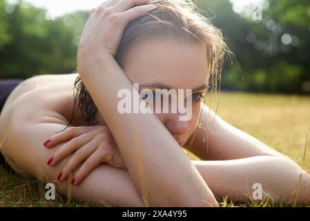 Close-up of a thoughtful woman lying on the grass on a sunny day, with her head resting on her arms. Stock Photo