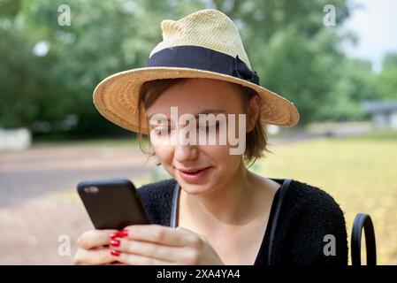 A smiling woman wearing a sun hat is looking at her smartphone in a park setting. Stock Photo