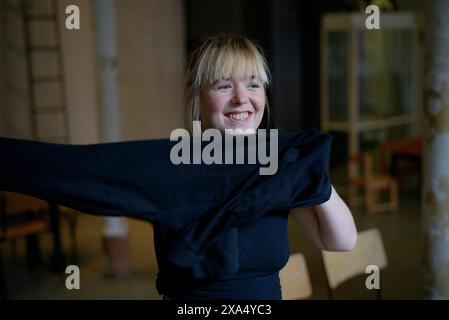 Smiling woman with blonde hair putting on a black shirt indoors. Stock Photo