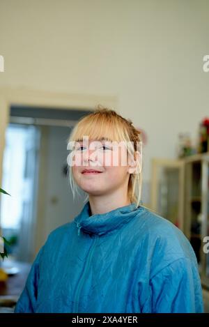 Smiling young woman with blonde hair in a blue jacket standing indoors. Stock Photo