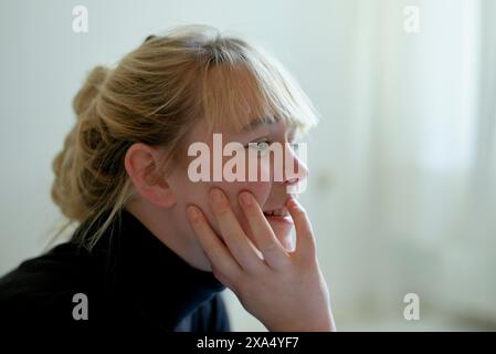 Smiling woman with blond hair in a messy bun resting her chin on her hand, looking away thoughtfully. Stock Photo
