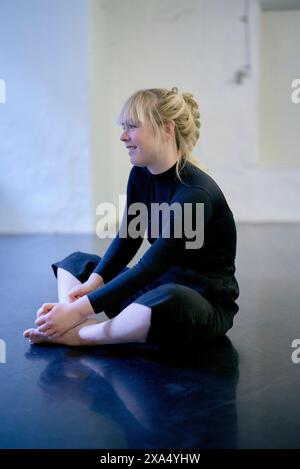 A smiling young woman in a black outfit sitting on the floor while stretching, with a light background. Stock Photo