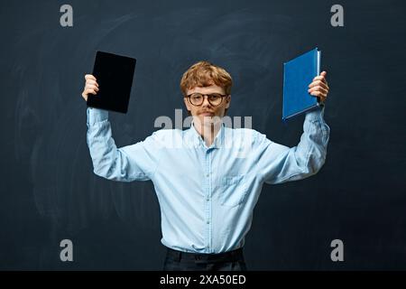 Young man with red hair and glasses stands holding tablet in his right hand and blue book in his left against writing board background. Stock Photo