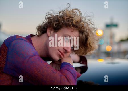Curly-haired young man leaning on a car at dusk with city lights in the background. Stock Photo
