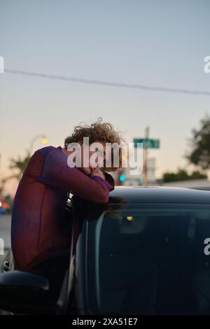 Young curly-haired man leaning on a car at dusk with city lights in the background. Stock Photo