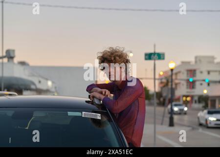 Curly-haired man leaning on a car roof at dusk in an urban setting. Stock Photo