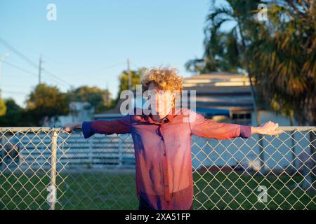 Young adult with curly hair leaning on a metal fence during sunset in a residential area. Stock Photo