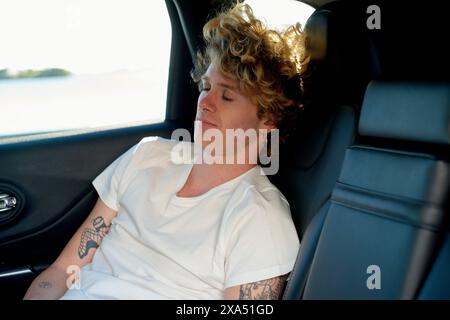 Young adult man with curly hair and tattoos sleeping in the back seat of a car. Stock Photo