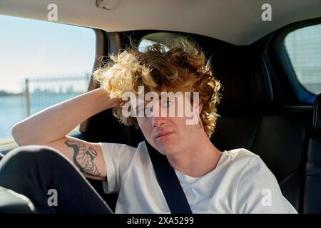 Curly-haired young man sitting in a car with a bridge in the background, looking pensively at the camera. Stock Photo