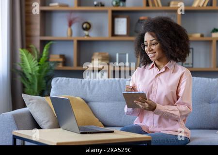 Young student joyfully learning from home, taking notes while engaged in an online video course on laptop. Comfortable home study environment. Stock Photo