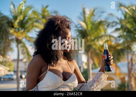 Young woman enjoying a soda bottle outdoors on a sunny day, with palm trees in the background. Stock Photo