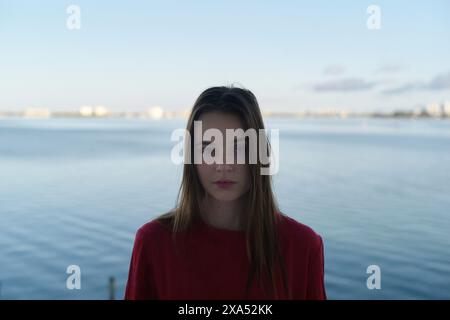Serene young woman in a red top with a waterfront view in the background. Stock Photo