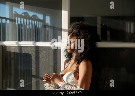 Serene young woman with curly hair enjoys the sunlight by a window. Stock Photo