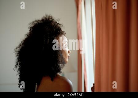 Young woman with curly hair looking out a window with a serene expression. Stock Photo