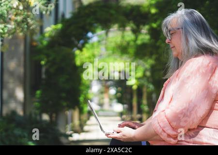 An older woman with white hair and glasses sitting on a park bench working with her laptop computer Stock Photo