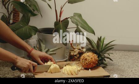 A person is slicing a pineapple on a cutting board next to a potted indoor plant in the kitchen counter, relaxing candid daily life at home Stock Photo