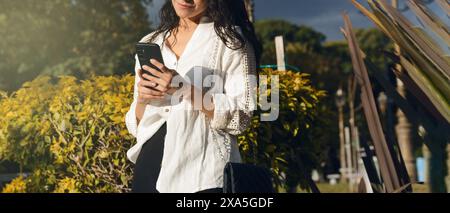 Banner of an unrecognizable young woman using the phone standing in a public park, is illuminated by the sunshine, an is holding her mobile phone with Stock Photo