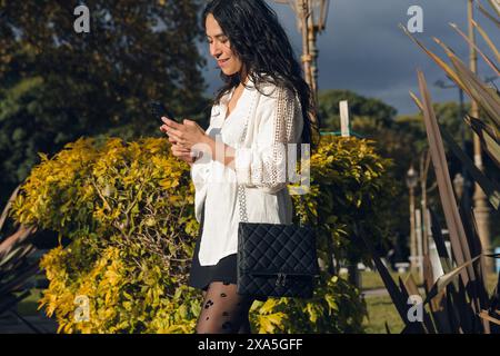 An Young Latin Woman is happy reading a message in her mobile phone standing in the park. She is smiling and she is enjoying her time outdoors. Stock Photo