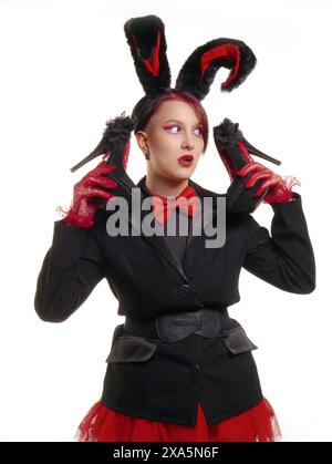 Stylish woman in bunny ears holding toy carrot and smiling on blue ...
