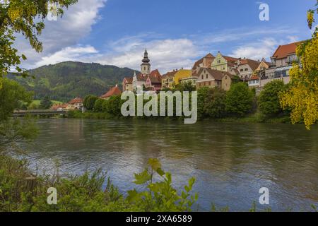 panoramic photo of a medieval Austrian village across Mur river on summer sunny day, pastoral Alpine mountains landscape Stock Photo