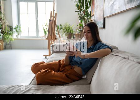 Peaceful pleased woman using smartphone, procrastinating relaxing resting on couch at home workshop Stock Photo
