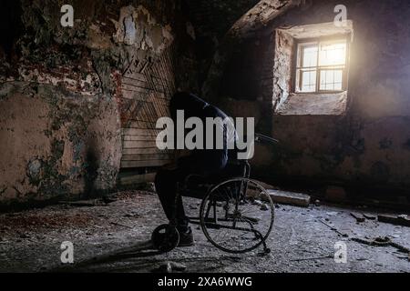 Sad man sitting in old wheelchair in abandoned hospital ward. Person silhouette in decayed vehicle for disabled patients in old building. Stock Photo