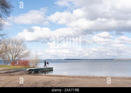 A Man and woman eagerly waiting for summer standing at end of dock on a sunny spring day with soft puffy clouds over the lake Stock Photo