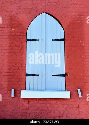 The closed blue wooden shutters of a window set in a bright red brick wall of Fisgard Lighthouse, Victoria, BC. Stock Photo