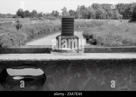 A monochrome close-up of a sizable bolt and nut on an aged bridge, against a river backdrop Stock Photo