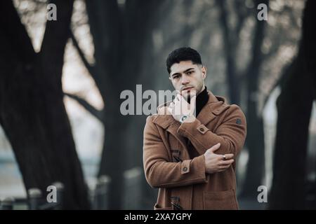 A man in a brown coat standing with crossed arms near trees Stock Photo