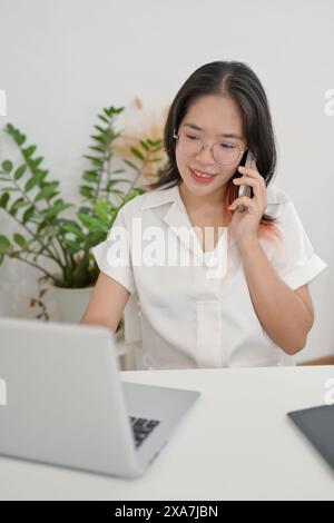 A young positive Asian woman is working remotely from a cafe, talking on the phone with her client while working on her laptop computer. multitasking, Stock Photo
