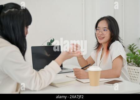 A young cheerful and friendly Asian female college student is meeting her friend at a cafe to work on a project together, talking, discussing on the p Stock Photo