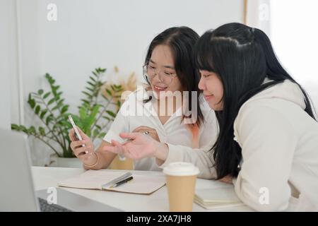 Two young, cheerful Asian female college students enjoy watching a video on a smartphone together while doing homework in a cafe. people and lifestyle Stock Photo