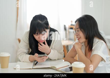 A young Asian woman in glasses is sitting with her friend in a cafe, studying or doing homework, while her friend searches for the answer on the inter Stock Photo