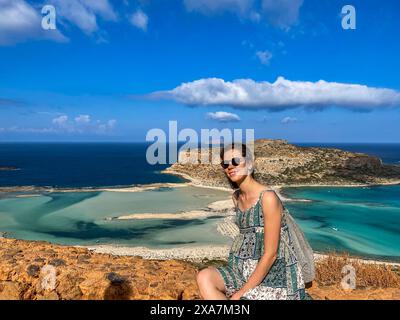 A woman sits on mountain edge, gazes at beach below Stock Photo