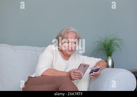 Older woman smiles while shopping on line using cell phone and card bank Stock Photo