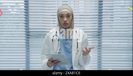 Image of young biracial female medical professional wearing hijab holding a tablet. she is standing in a modern medical office with blinds covering wi Stock Photo